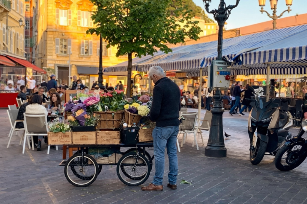 Kukkamyyjä torilla Nizzassa ilta-auringossa, taustalla ravintoloita ja vilkasta katuelämää