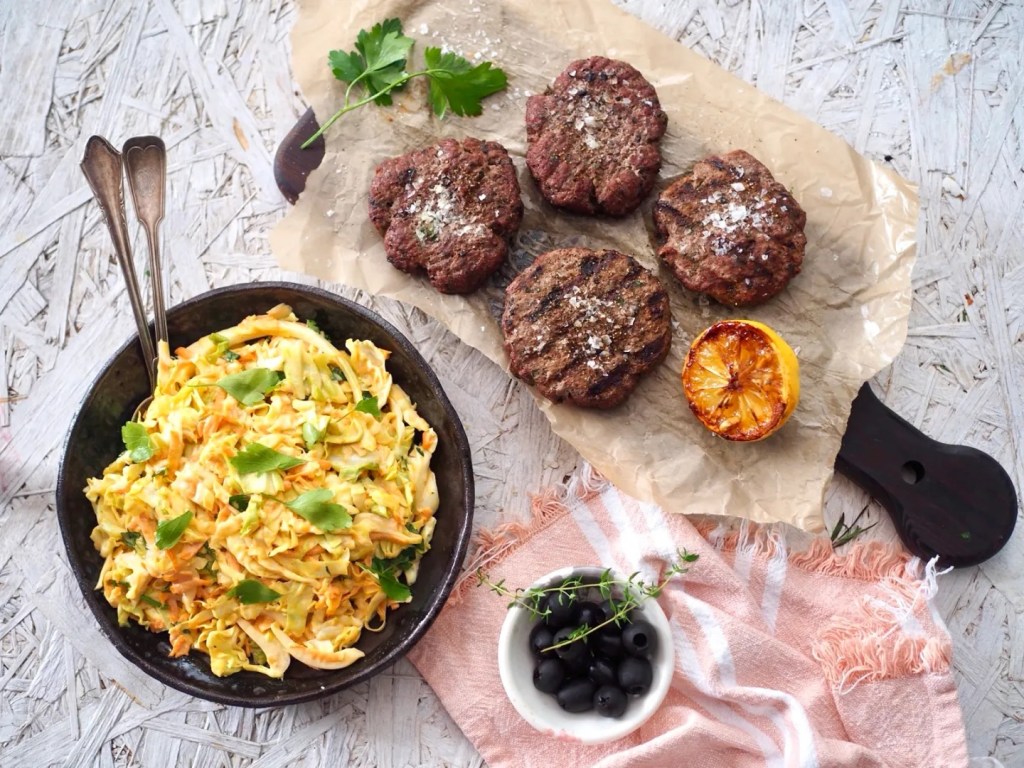 Karitsanlihapurilaiset ja varhaiskaali-coleslaw tarjoiltuna kulhossa, vierellä grillatut pihvit ja sitruuna.