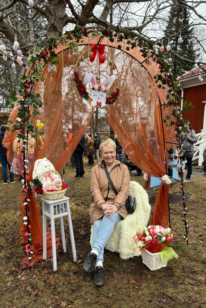Nainen istuu pääsiäiskoristellun kaaren alla Öllerin talon pihalla Riihimäellä, kukkia, pääsiäispupu ja keväinen tapahtumatunnelma