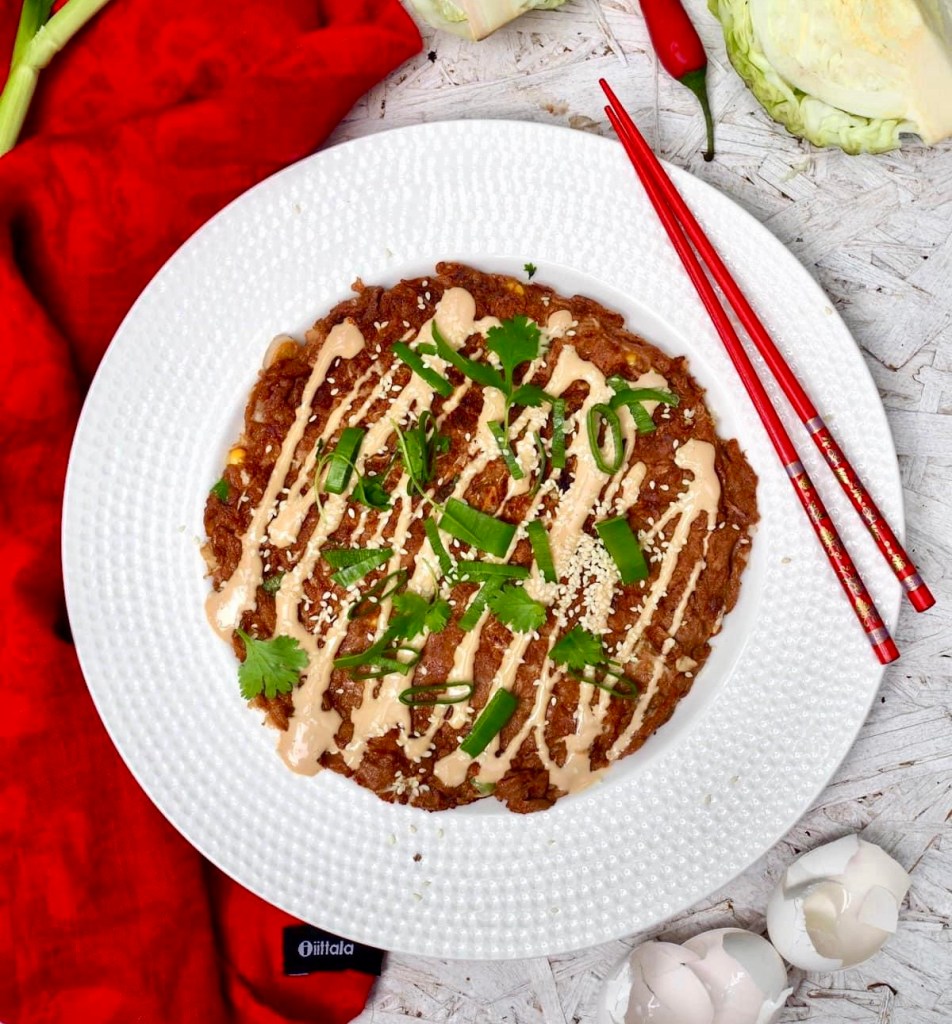 Okonomiyaki eli japanilainen kaalimunakas lautasella, päällä kastiketta, seesaminsiemeniä ja kevätsipulia.