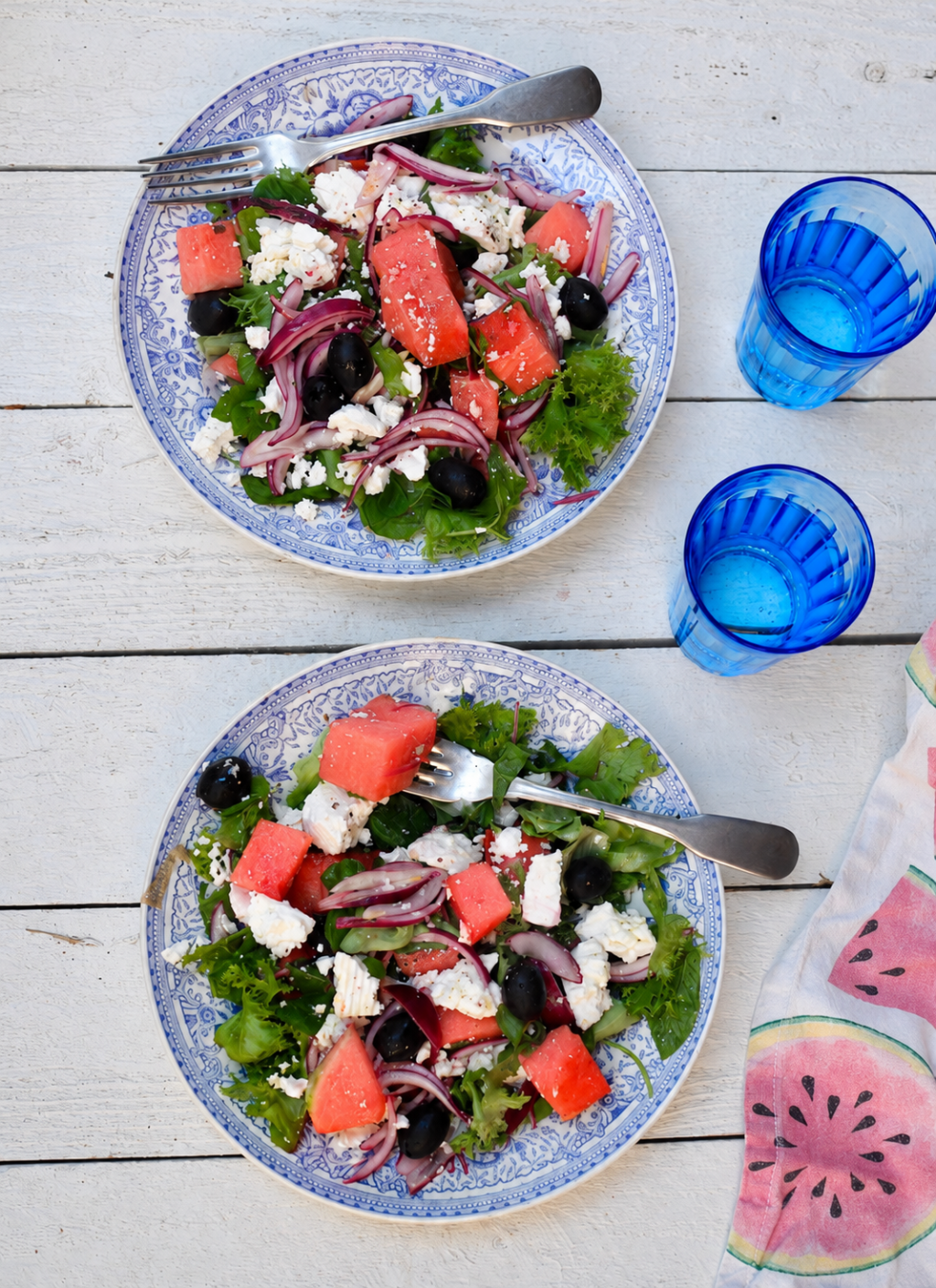 Vesimeloni-feta-oliivisalaatti kahdella sinivalkoisella lautasella valkoisella puupöydällä. Salaatissa on vesimelonia, fetajuustoa, mustia oliiveja, punasipulia, vihersalaattia ja tuoreita yrttejä.
